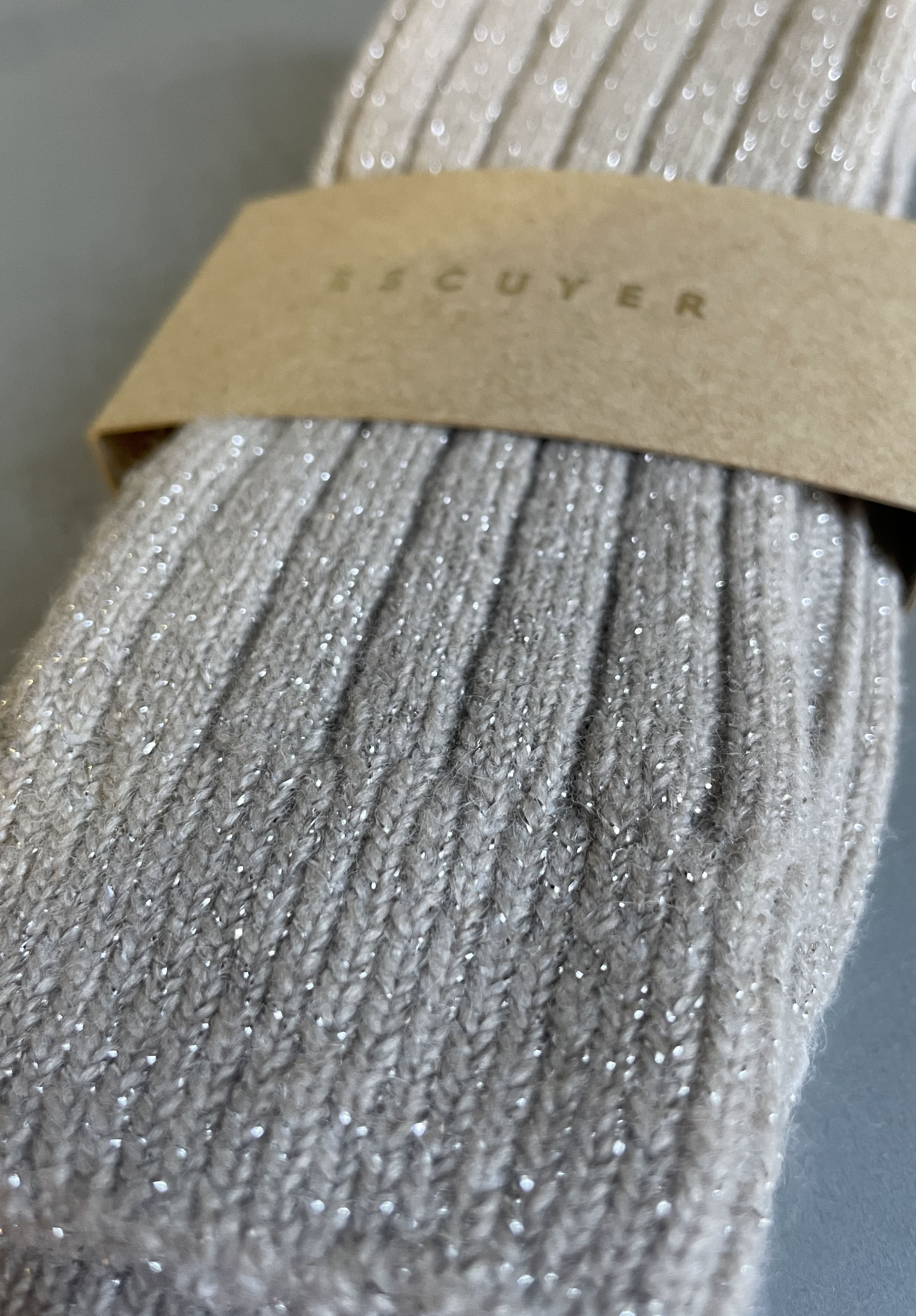 A close-up of a ribbed knit sock in a light taupe-gray color with subtle silver glitter, wrapped in a brown kraft paper band printed with the brand name ESCUYER.