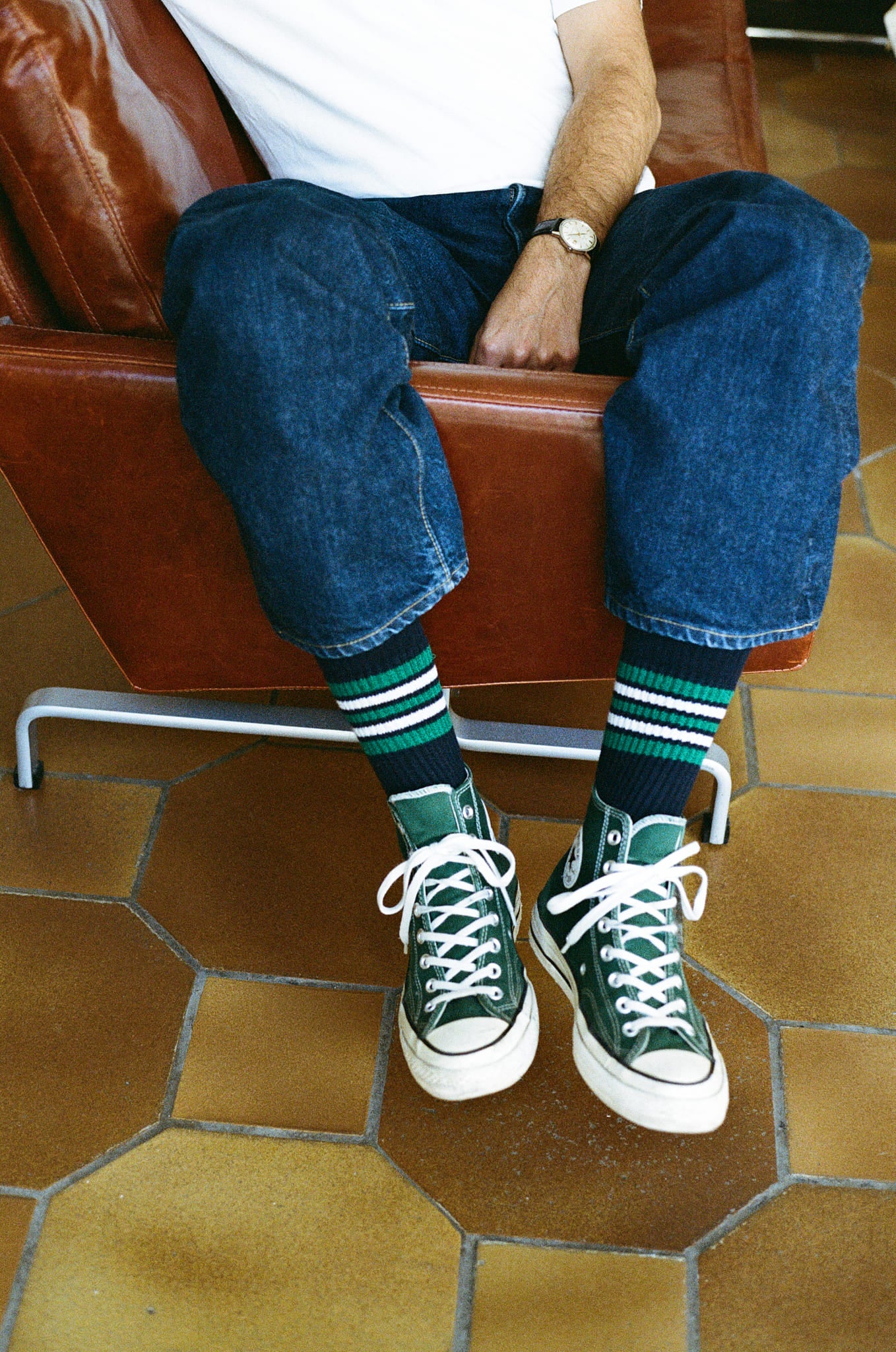 Person wearing green sneakers and blue jeans sitting on a brown leather chair with a tiled floor.
