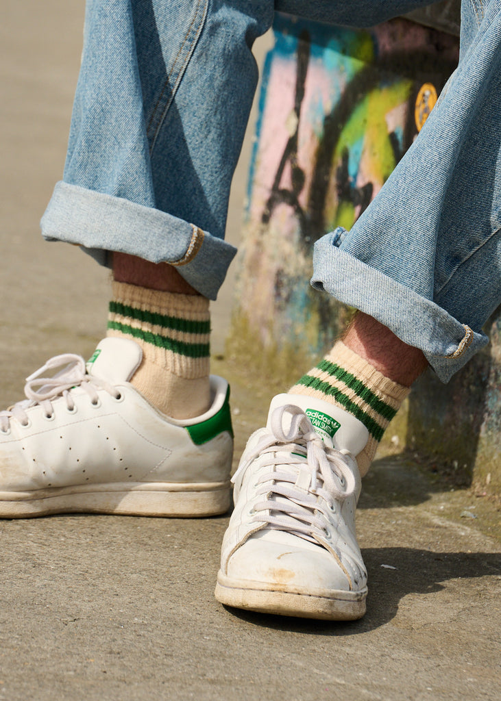 Close-up of feet wearing white sneakers with green stripes, beige socks, and rolled-up jeans against a blurred background.